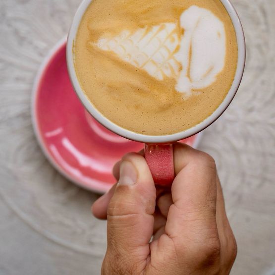 Kávová zmrzlina k Mezinárodnímu dni kávy Ruka držící růžový šálek kávy s latte artem ve tvaru zmrzlinového kornoutu.